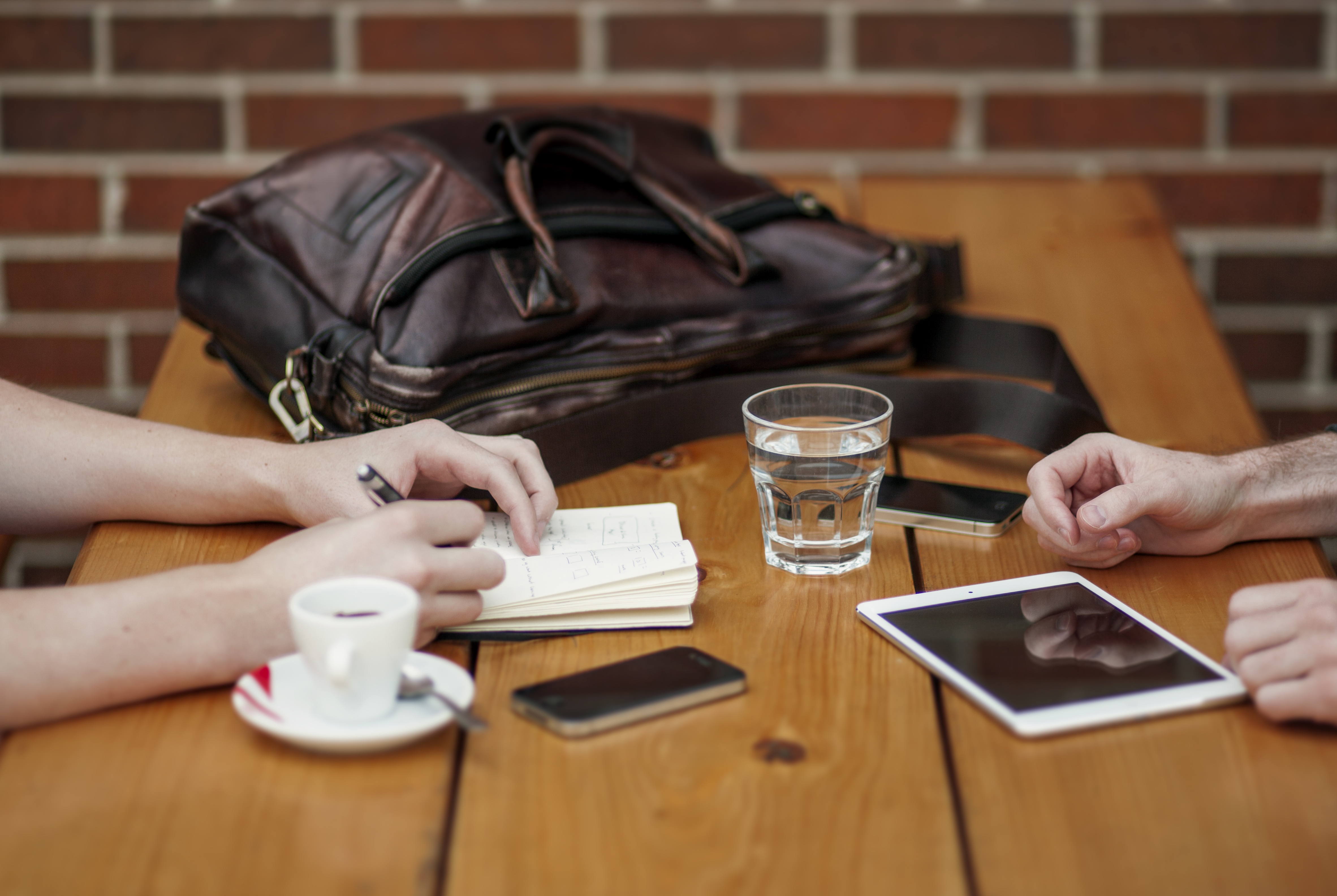 A bag lying on a desk with a hand writing on a notepad, a coffee cup and a glass of water next to it